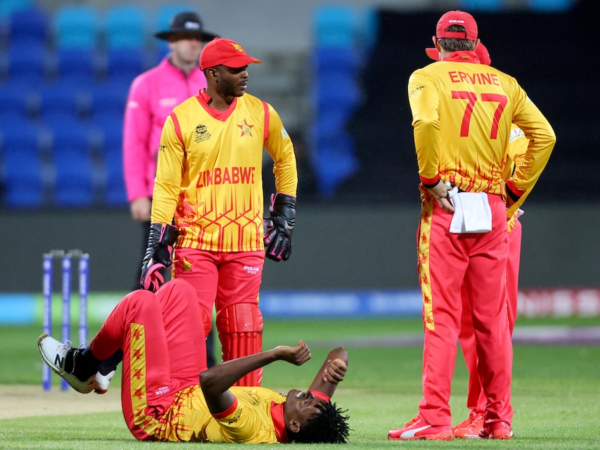 A Zimbabwe bowler lies on the ground in pain while teammates stand around during a cricket match.