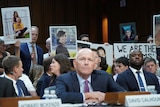 People hold photographs and behind man sitting in hearing. 