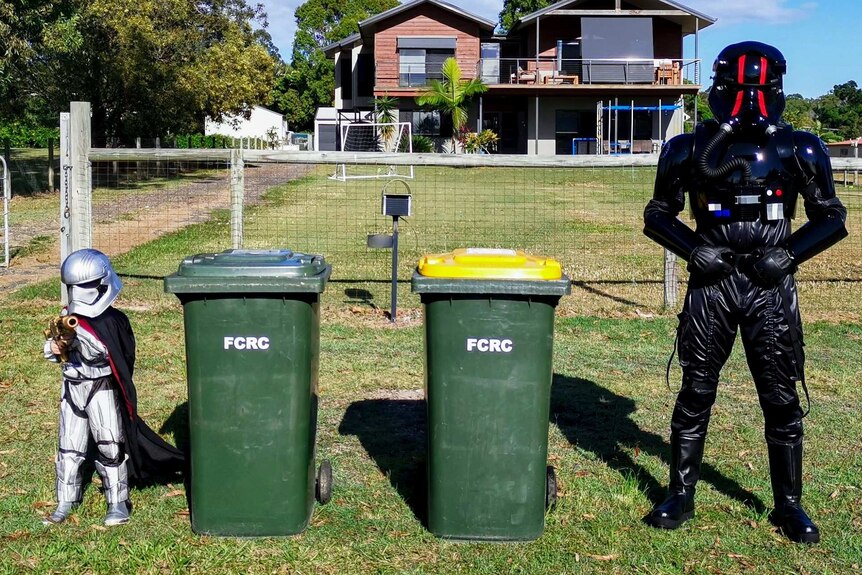 A small child in a Captain Phasma outfit and an adult in a Tie pilot costume standing next to two kerbside bins.