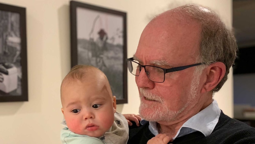 An older man with a beard holding a baby in a hallway