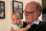 An older man with a beard holding a baby in a hallway