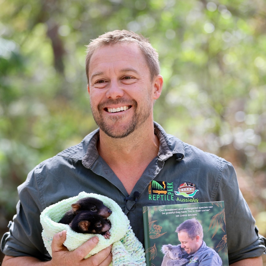 A man, holding a Tasmanian Devil and a book, looks off camera smiling