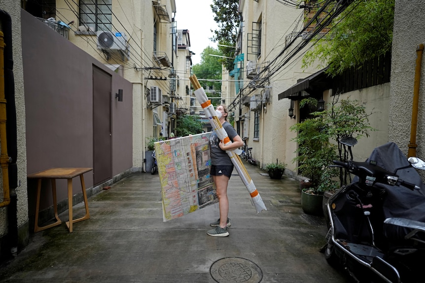 A woman in shorts and T-shirt carries long rods and a large artwork as she stands in an alleyway
