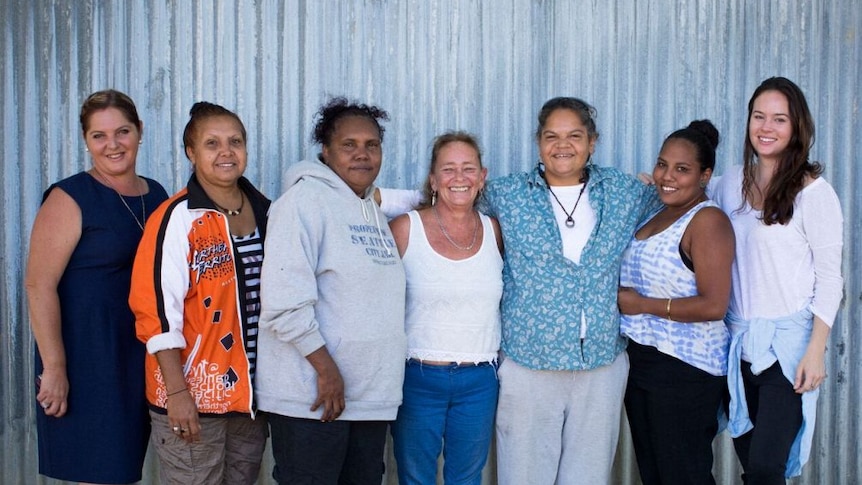 seven women standing in front of a corrugated iron shed