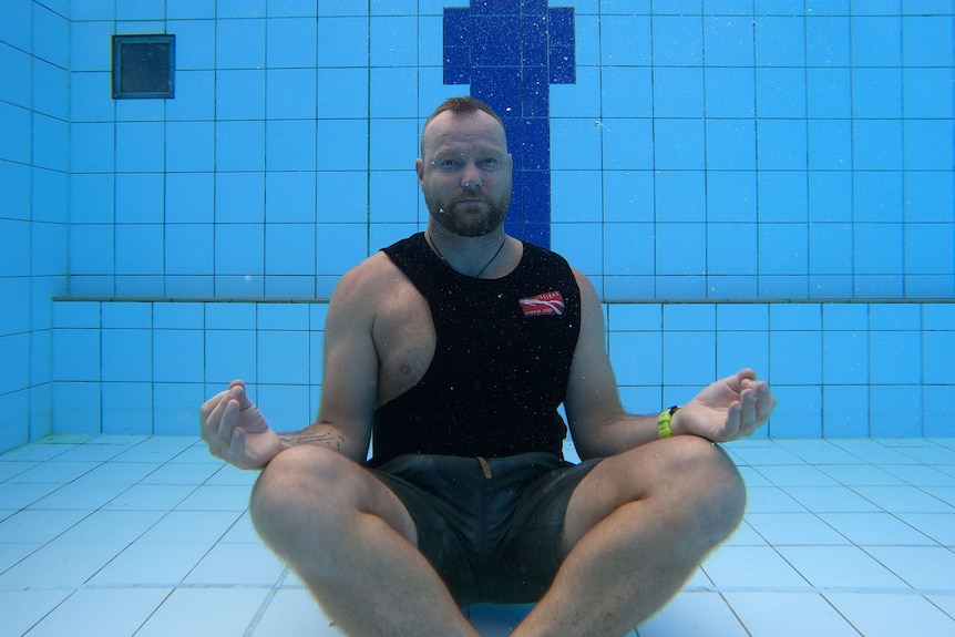 A man with short hair and a neat beard sits in the lotus position on the bottom of a pool.