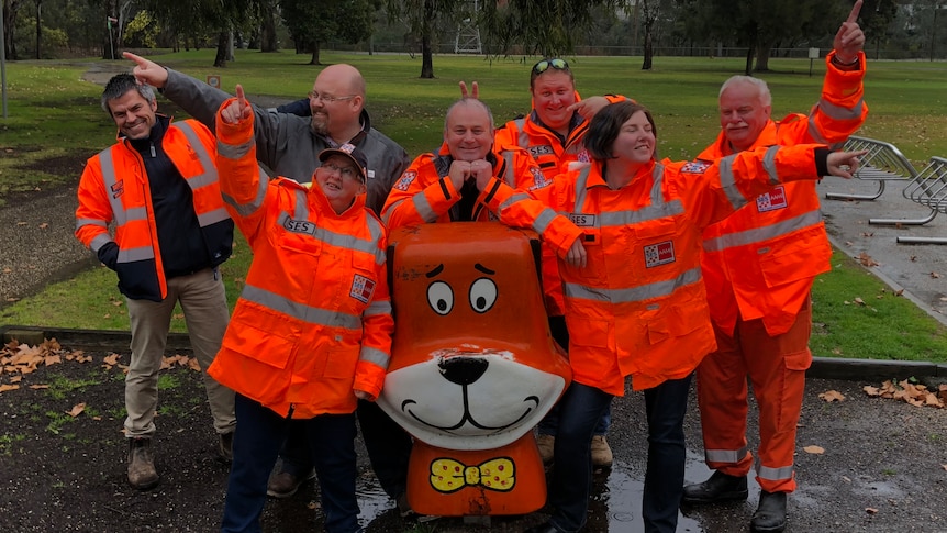 Seven volunteers from the Wangaratta SES Unit pose alongside the Yogi statue in their fluoro orange vests.
