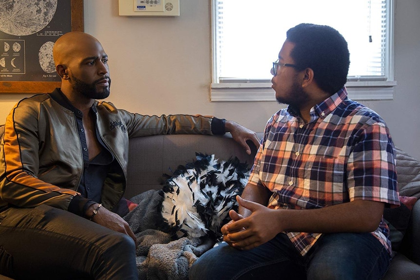 Karamo Brown sits on a couch listening as another man speaks