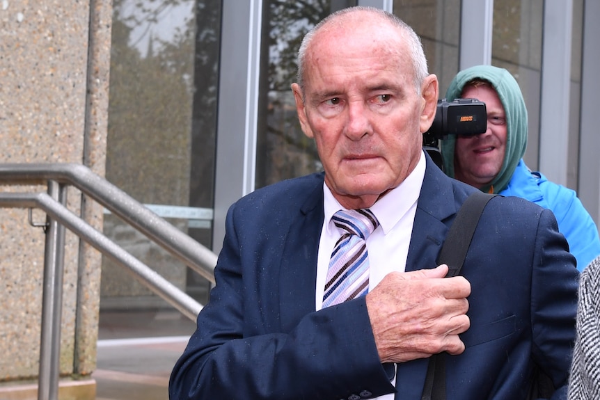 A stern man in a suit walks outside court as a video photographer follows him