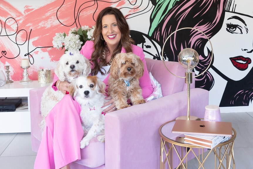 A women in a pink suits, sits on a pink armchair, holding three dogs. 