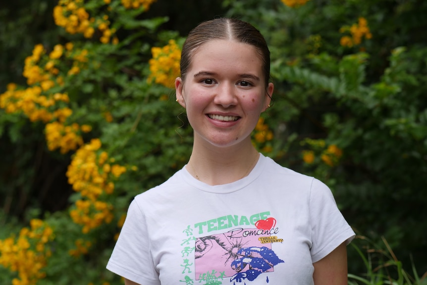 Teenage girl looks at the camera smiling. Her hair is tied back and she is wearing a t-shirt with the words 