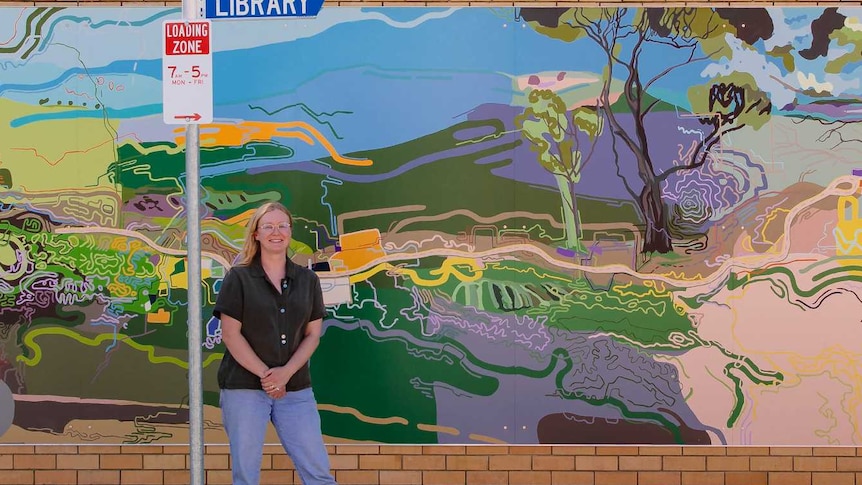 A blonde-haired lady wearing a black shirt and jeans wearing glasses stands next to a street sign in front of a mural with lines