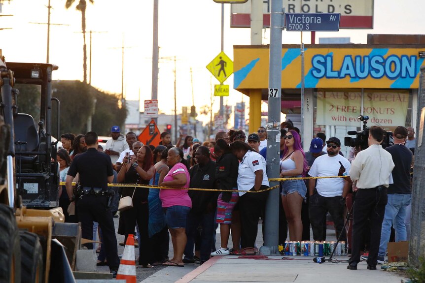 A crowd of people gather close to a crime scene on a road lined with shops.