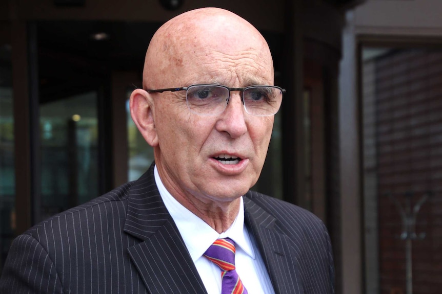 A mid-shot of WA Attorney-General John Quigley wearing a suit and tie speaking to unseen reporters.