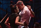 On stage, a Latino woman stands close to a muscular man with a buzz cut. She is holding him at his sides.