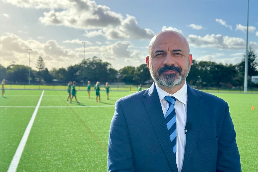 A man in a suit with a beard stands on a soccer pitch and smiles into the camera.