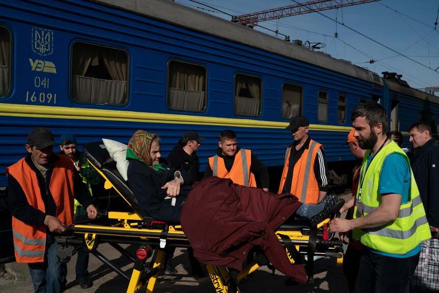 An elderly woman lies on a wheeled stretcher being pulled along by a group of men in high-viz vests, next to a train.