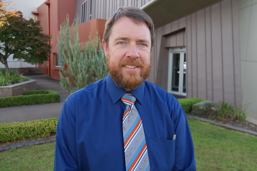 A man with red hair and a beard smiling in front of a building and a bush, wearing a blue shirt and a colourful tie.
