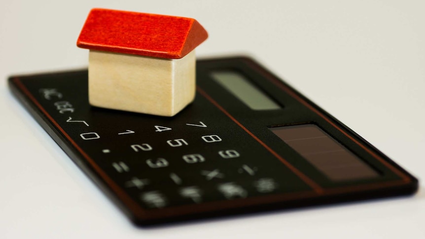 A small wooden toy house with a red roof sits on top of a thin black calculator on a white surface.