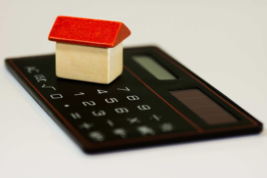 A small wooden toy house with a red roof sits on top of a thin black calculator on a white surface.