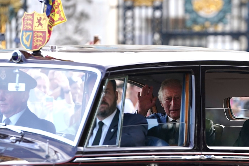 Charles waves from the backseat of a car, driven by a royal driver.