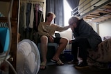 A small room with a teenage boy on a chair and a blonde woman on the side of a bunk.