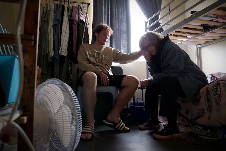 A small room with a teenage boy on a chair and a blonde woman on the side of a bunk.