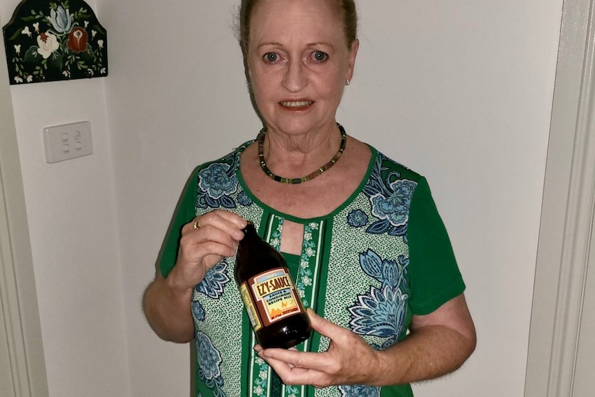 A woman with her hair in a bun, wearing a green necklace and top holds up a brown bottle of EZY Sauce