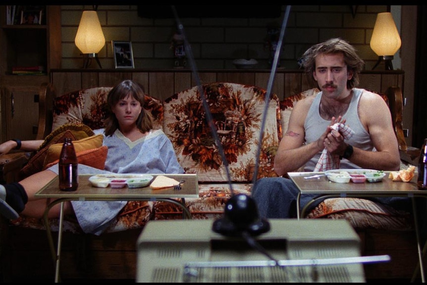 Colour still of Holly Hunter and Nicholas Cage sitting in front of television in lounge room in 1987 film Raising Arizona.