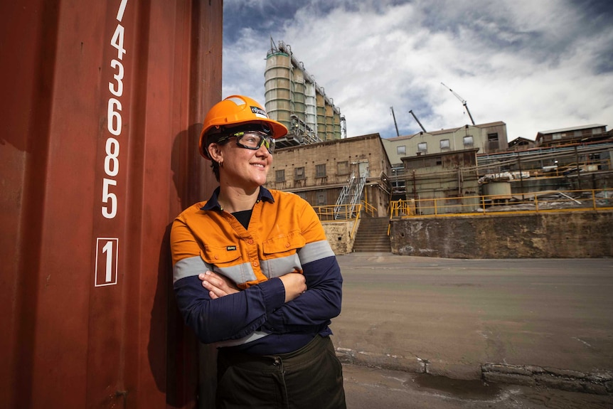 A woman in an orange hard and overalls hard looks to the right while standing at a zinc smelter