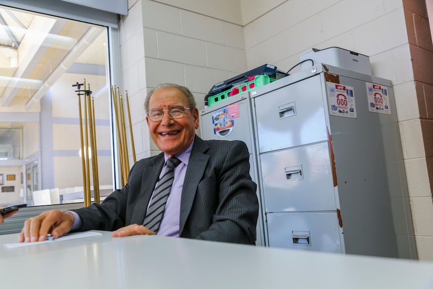 A man smiles sitting at a meeting room desk. There is a filing cabinet behind him