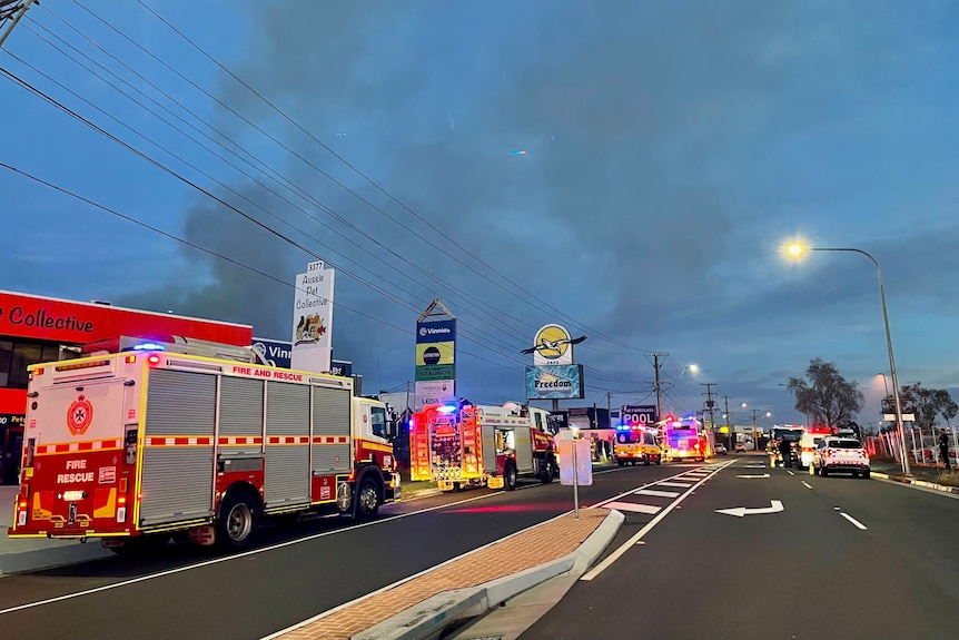 Smoke billows into the morning air on a busy commercial road. Fire trucks line the curb. 