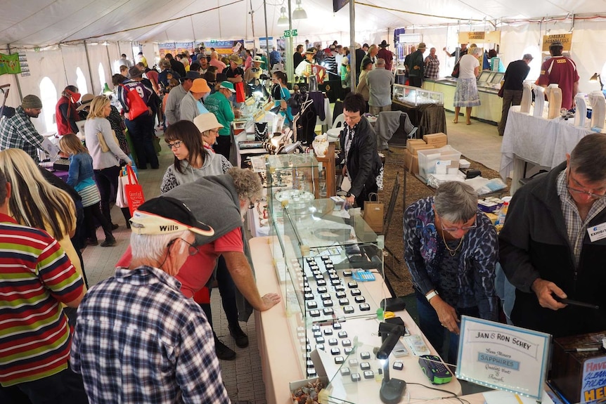 People stand in a large tent looking at market stalls showing gems and jewellery.