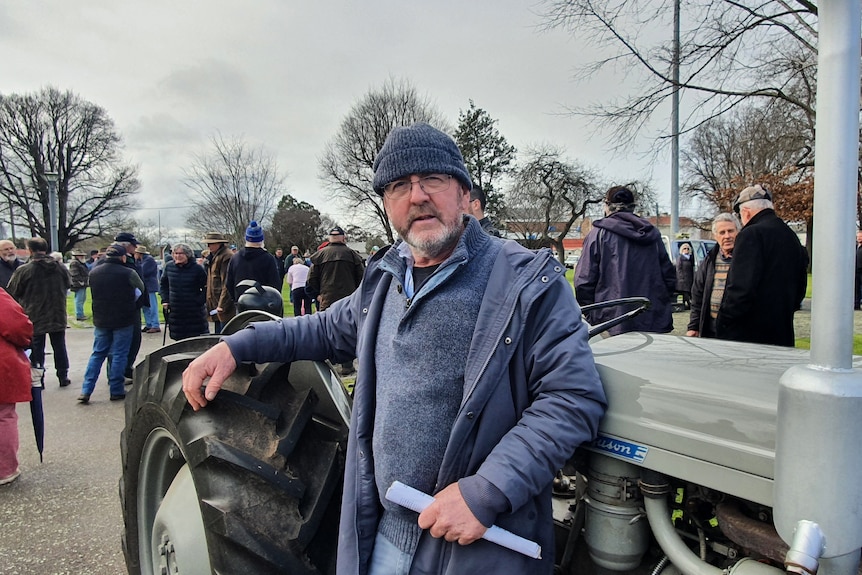 A bespectacled man wearing warm clothes leans on a tractor.