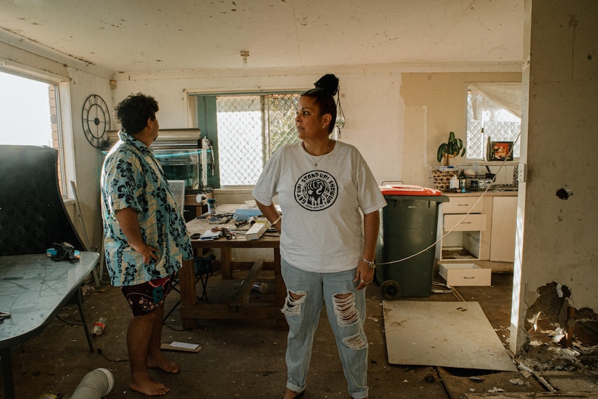 Two stand in the living room of a house, there is mess and debris, the floor and walls are damaged, a bin stands in the kitchen.