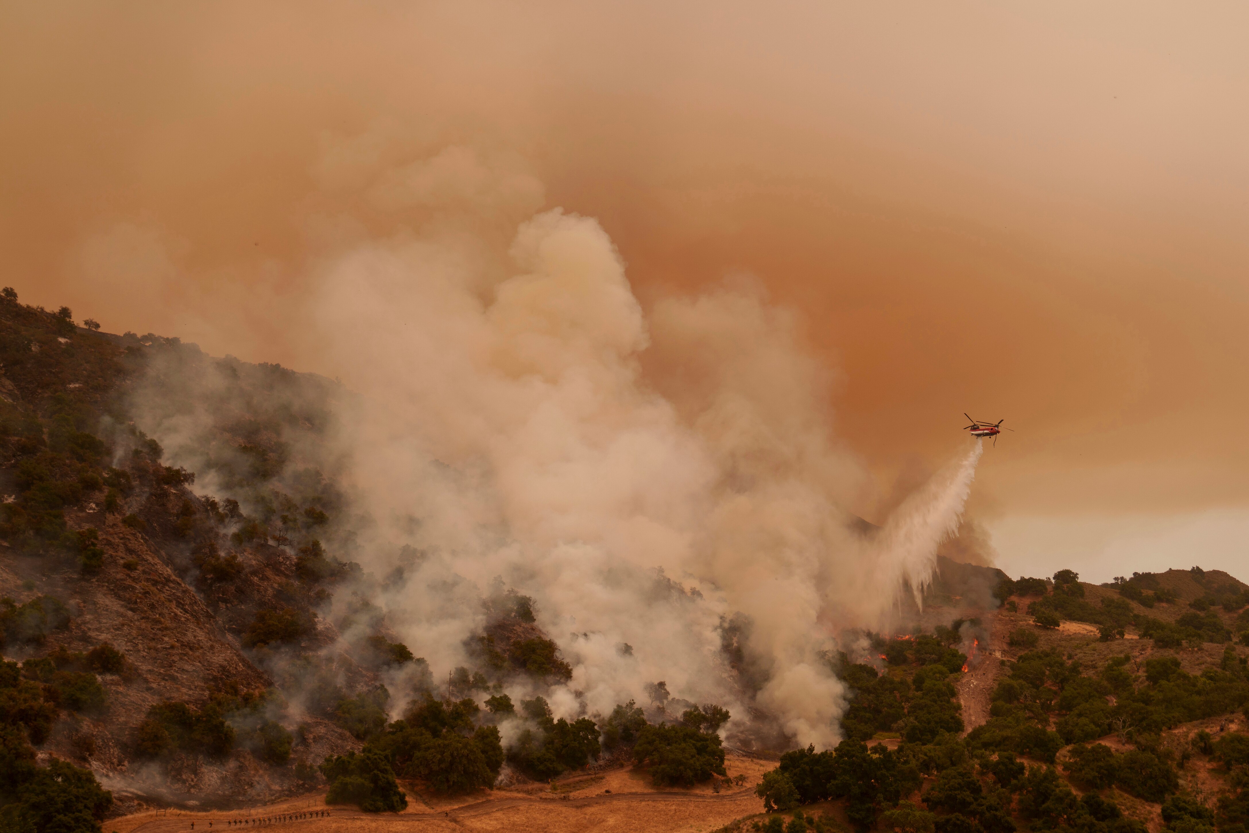 Smoke rises from an arid landscape, as a helicopter emerges on the right.