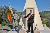 A man in a suit and sash stands in front of a monument with a man holding a flag and wearing army fatigues standing next to him.