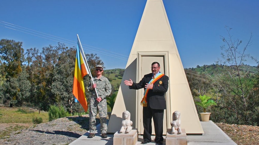 A man in a suit and sash stands in front of a monument with a man holding a flag and wearing army fatigues standing next to him.