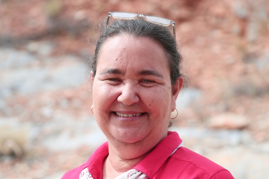 An Aboriginal woman in a red collared shirt smiles at the camera in a bush setting