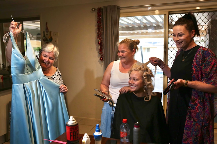 An older woman holds up a blue dress, while a younger woman sits in a chair having her hair styled.