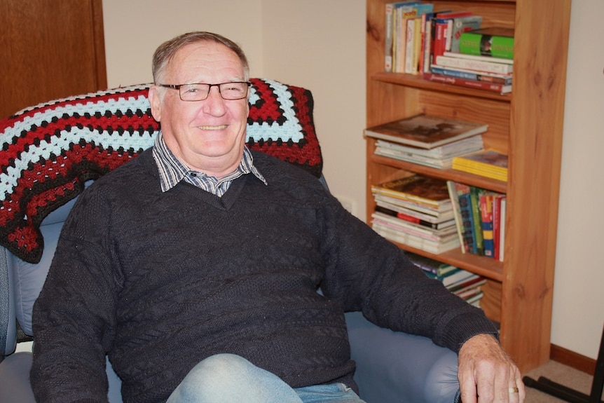 Laurie Allen, dementia carer, sits in armchair, smiles at camera. Dressed in grey jumper and chequered shirt.