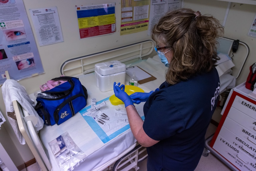 A nurse in scrubs and surgical mask prepares a vaccine vial for injection.