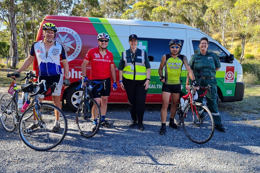 Three cyclists and two people in St John Ambulance uniforms stand in front of an ambulance and some bushland