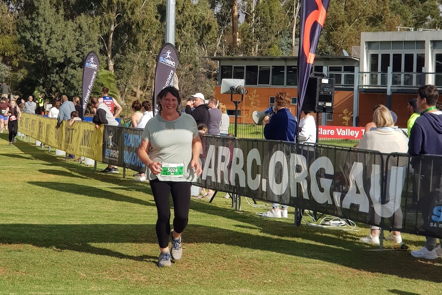 A woman in a blue tee and a race bib running and smiling below a finish line banner. 