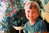 A little boy with short hair, a blue t-shirt and a beaming smile looks toward the camera. He sits in front of a garden. 