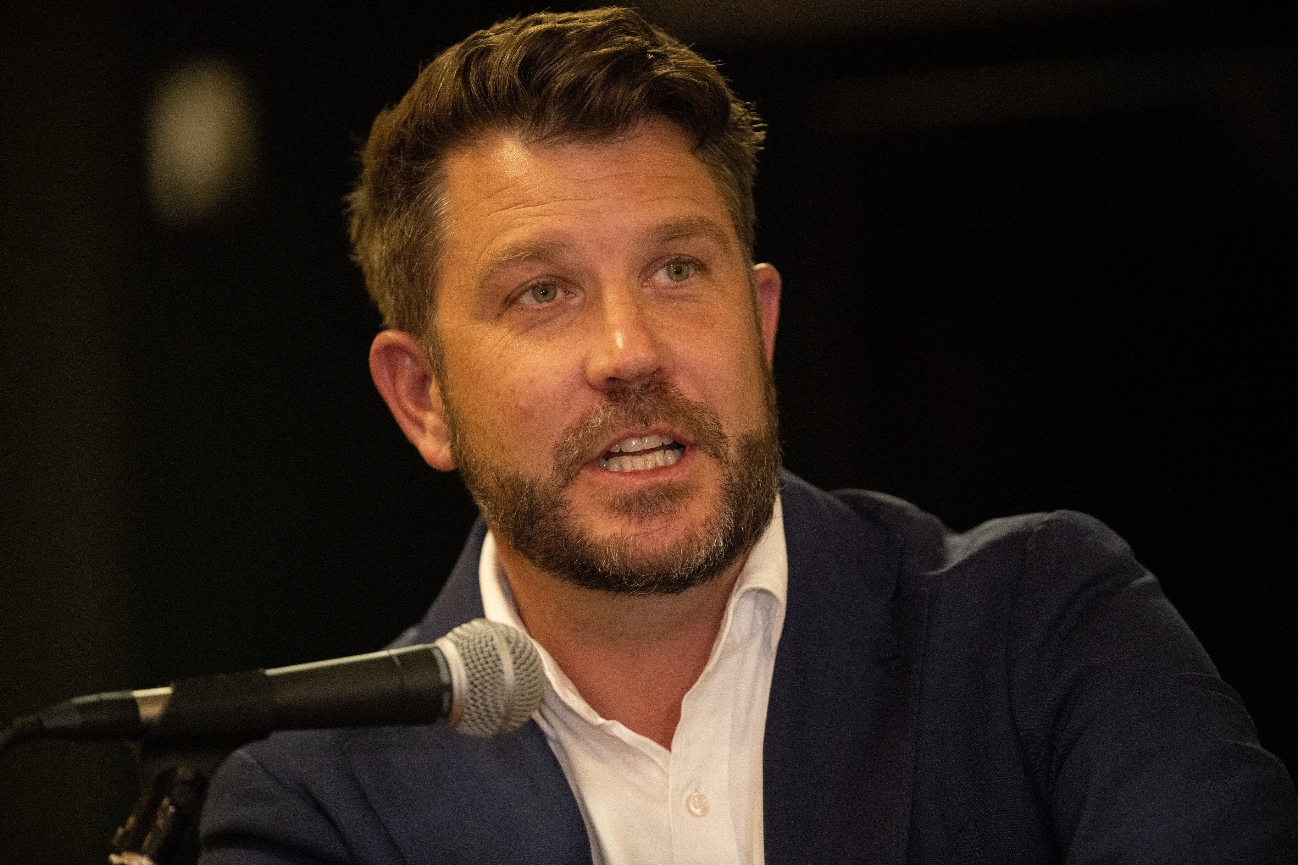 Close up photo of Moran speaking into a microphone, wearing white collared shirt with dark suit jacket 