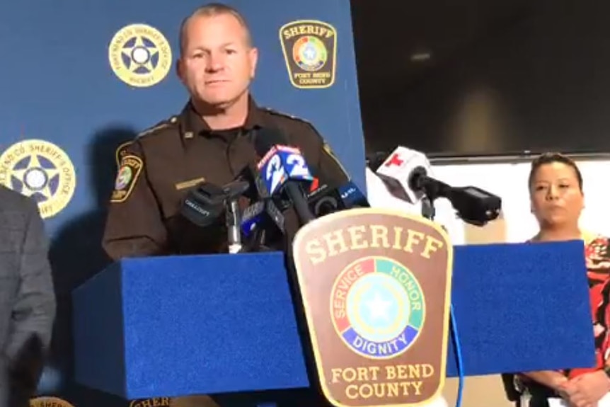 Troy Nels stands at a lectern with microphones, wearing a stern expression and a sheriff's uniform.