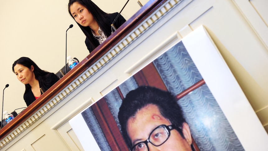 Two women give a press conference with a poster of a man displayed on the front of the bench.