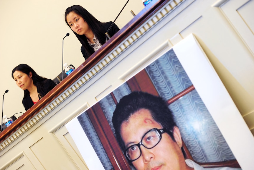 Two women give a press conference with a poster of a man displayed on the front of the bench.