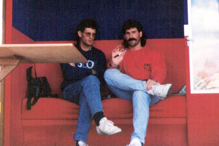 Two men sit on a large red couch in conversation. Their clothes are dated and the photo is from the 1980s