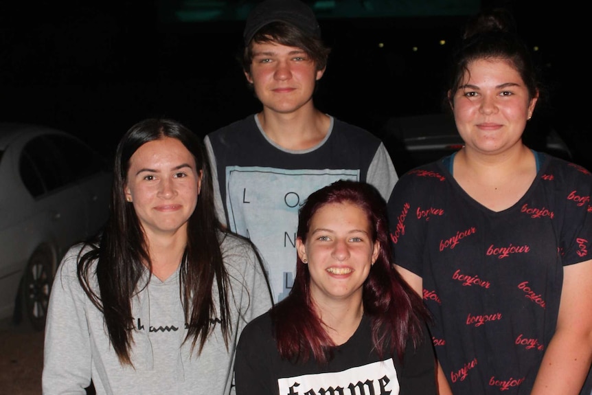 Four young adults stand in dim light with a projection screen looming large against a dark black background.